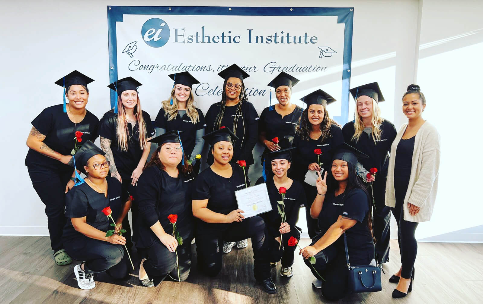 Group of graduates wearing caps and gowns holding roses, posing in front of a "Congratulations on your Graduation" banner at Esthetic Institute.