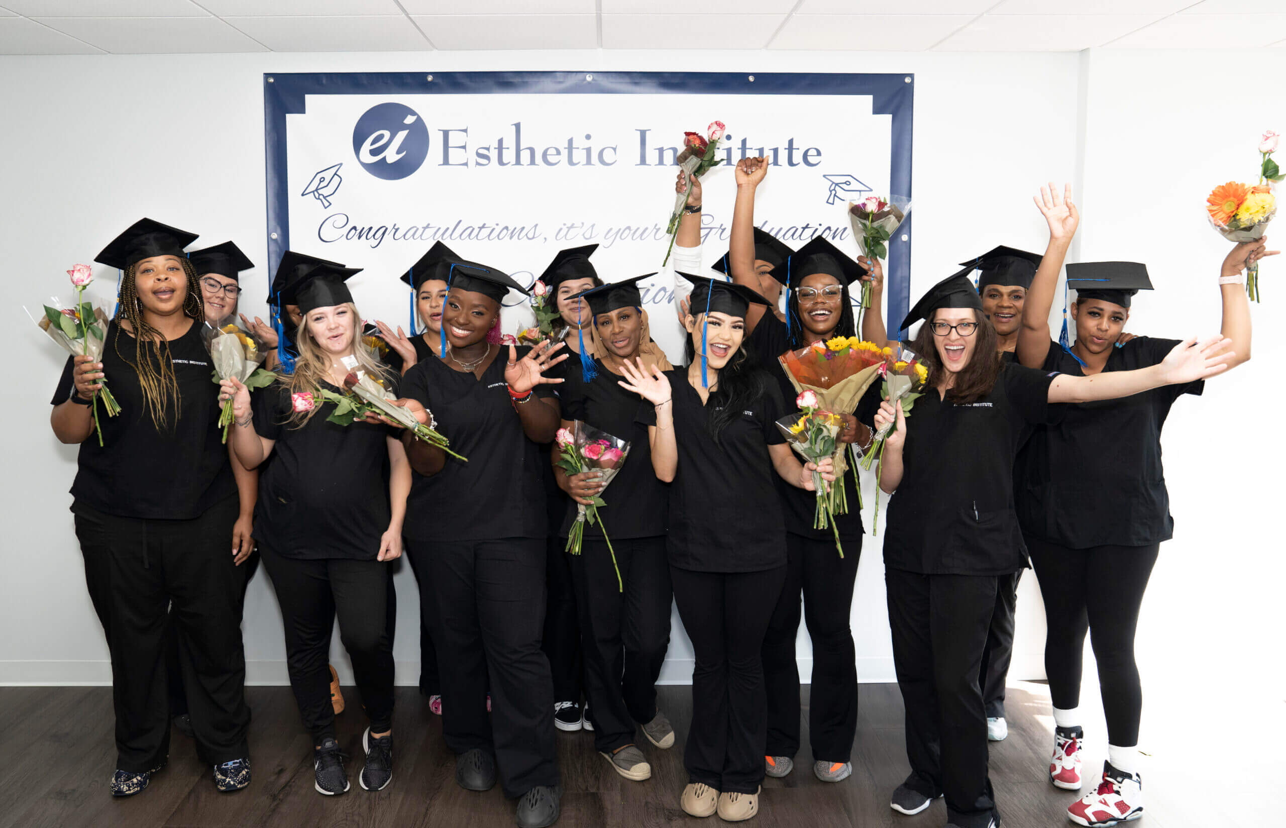 A group of people wearing graduation caps and black attire celebrates by holding flowers in front of a banner that reads "Esthetic Institute.