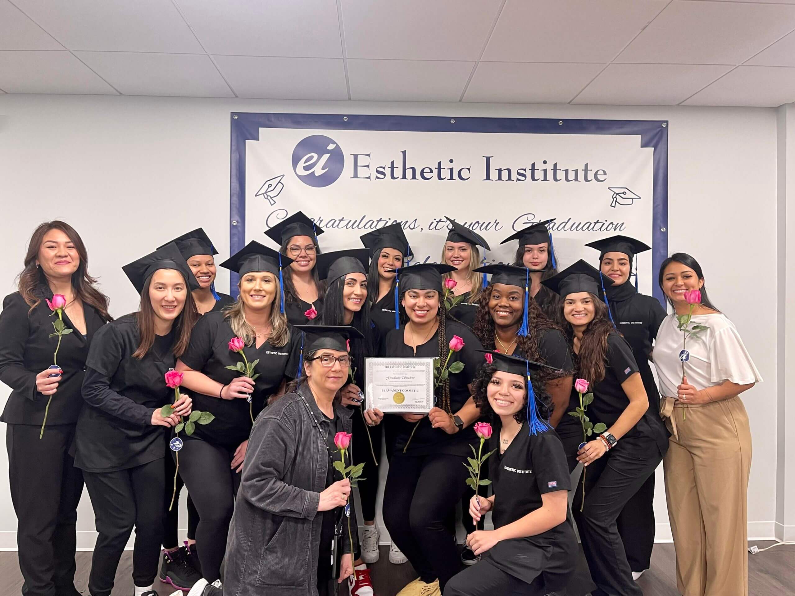 Group of graduates in caps and gowns holding roses, posing with a certificate in front of an "Esthetic Institute" banner.