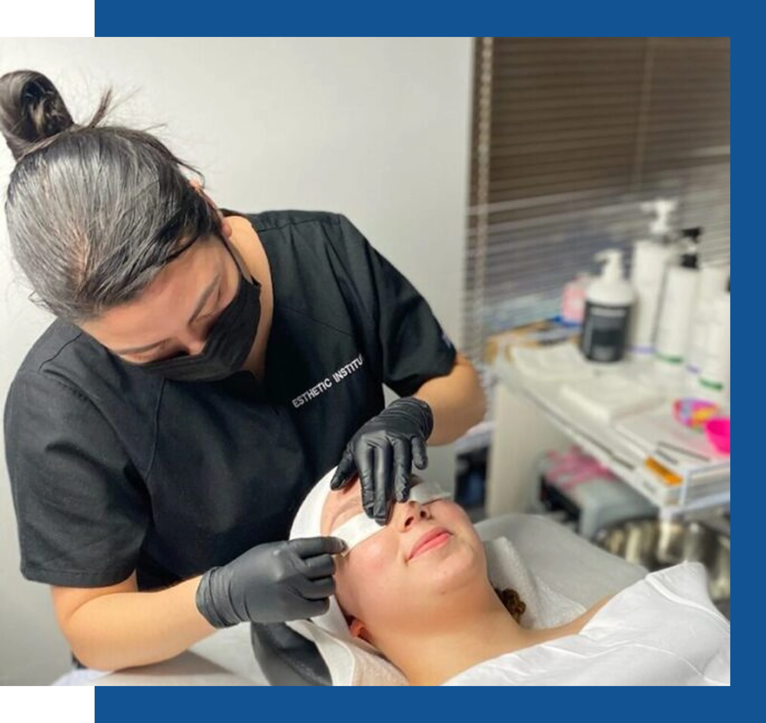 A skincare professional wearing gloves and a mask applies a treatment to a client lying on a bed in a clinic setting.