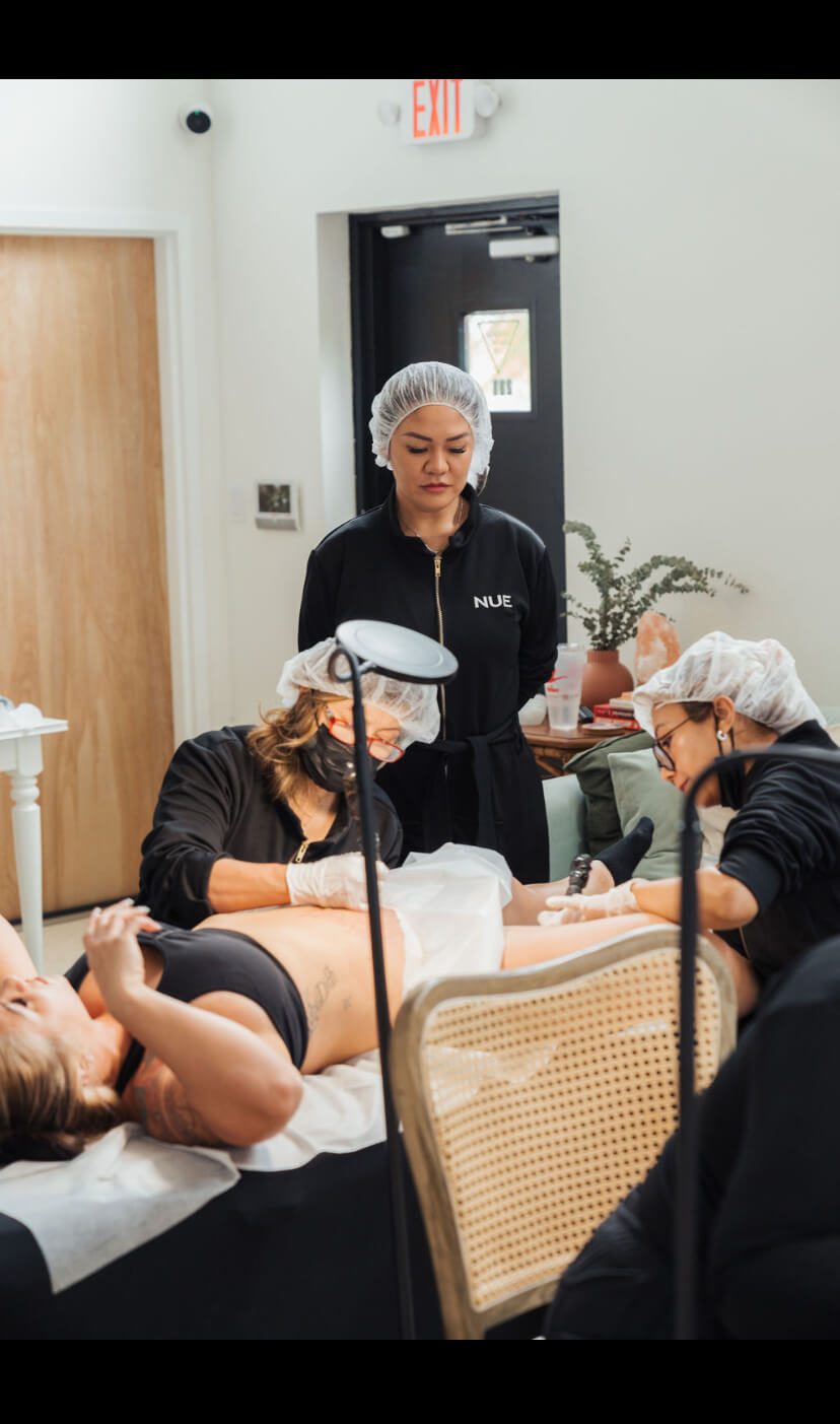 A woman lying down receives a cosmetic procedure from two practitioners, all wearing hair nets, in a clinical setting with a third person observing.
