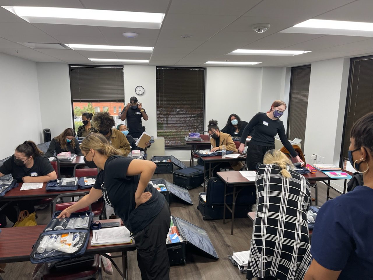 Room with people sorting medical supplies into cases. Several individuals are wearing masks and uniforms, while others are assisting. Tables and chairs are arranged around the room.