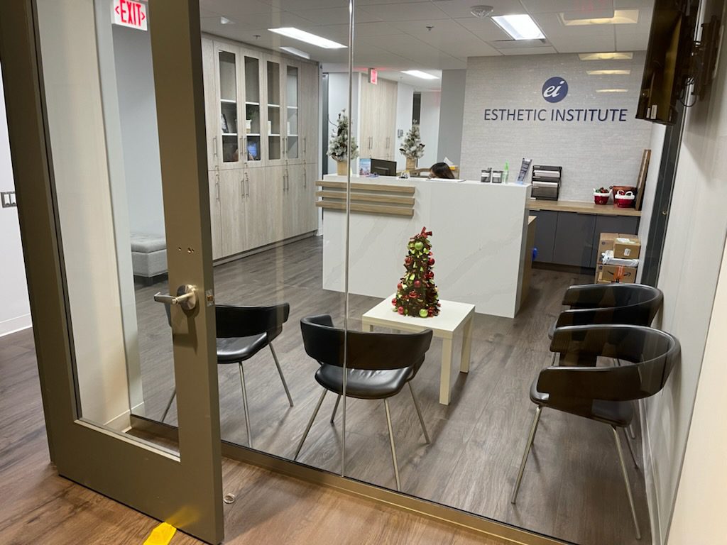 A small waiting area with black chairs and a table holding a mini Christmas tree. Behind glass doors, a reception desk with a computer is visible at the Esthetic Institute.