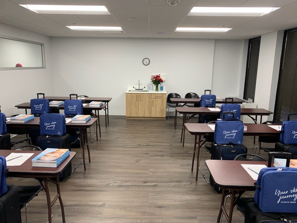 Classroom with rectangular tables and chairs, each setup with blue bags, books, and papers. A clock and flowers on a cabinet are visible at the back.