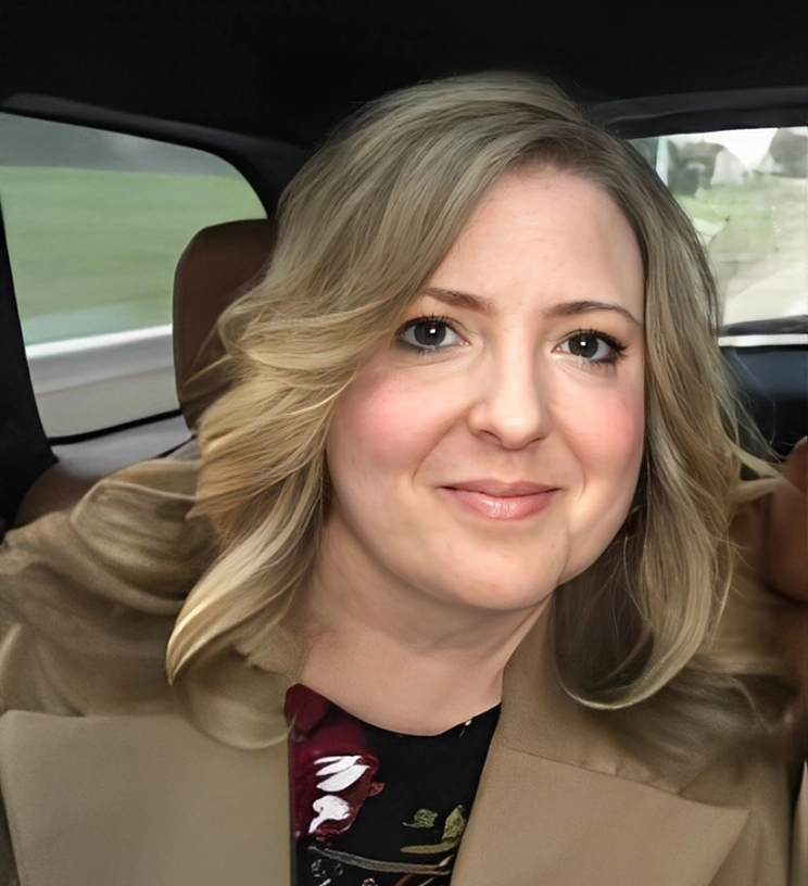 Person with shoulder-length blond hair, wearing a brown coat, sitting inside a car.