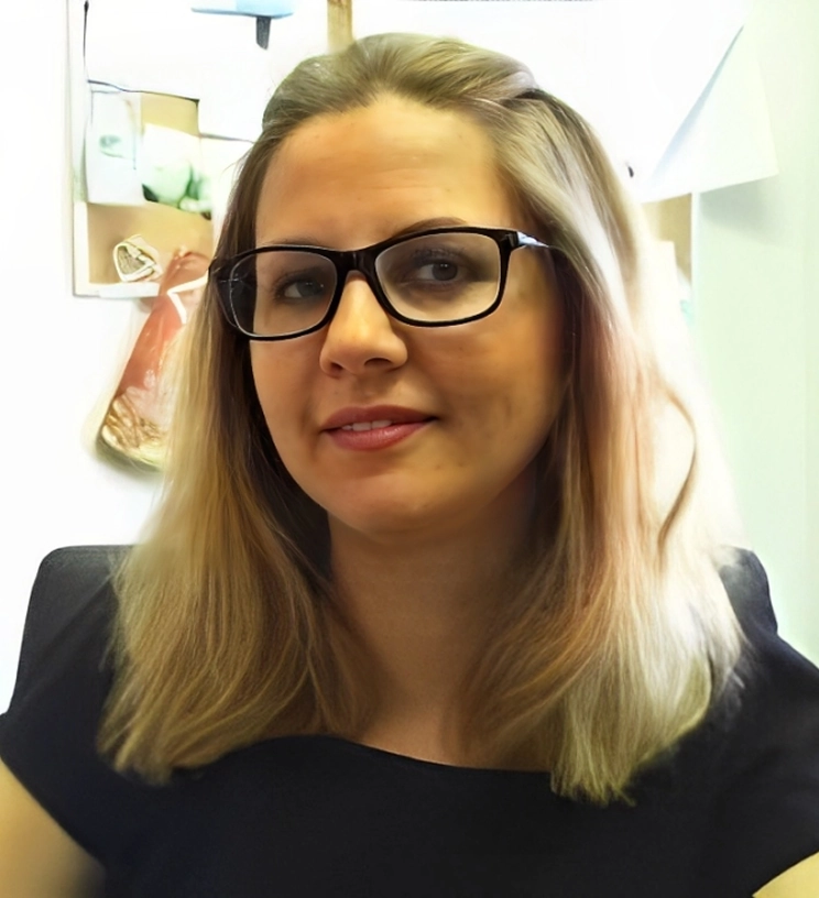 Woman with long blonde hair and glasses sitting in an office environment, wearing a black top, with a bulletin board visible in the background.