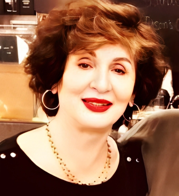 A person with short, wavy brown hair and red lipstick, wearing hoop earrings and a necklace, smiles at the camera.