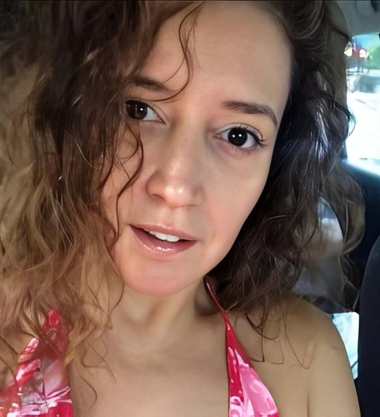 A person with curly hair wearing a red patterned top looks at the camera. The background is blurred.