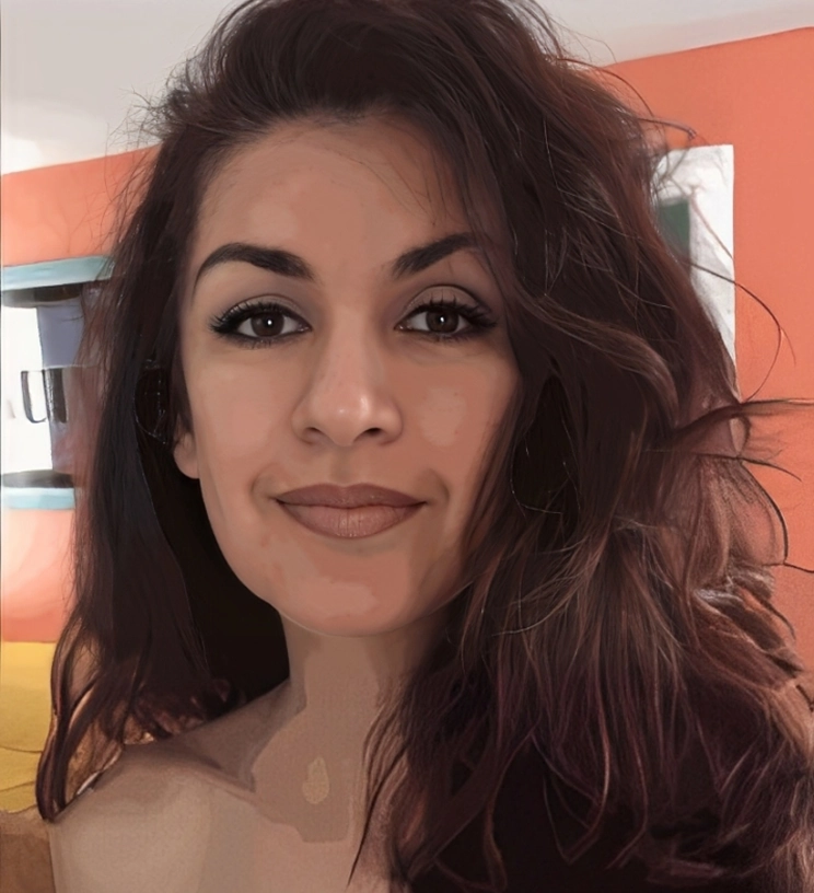 Woman with long wavy hair and subtle makeup, smiling slightly at the camera. Background features orange and white walls and a small part of a painting.