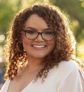 Mary-N-PMU-sm-2-5-274x300 (2) A woman with glasses and curly hair smiling.