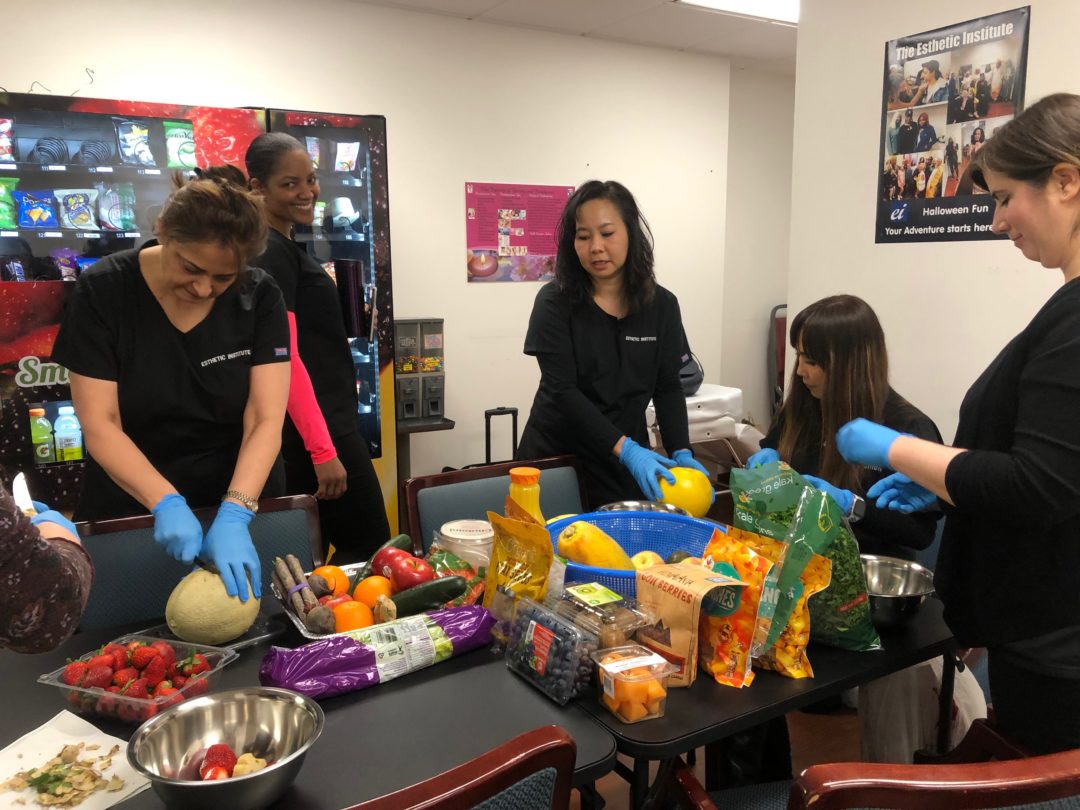 A group of people in black attire and blue gloves are preparing various fruits and vegetables on a table in a room with snack vending machines.