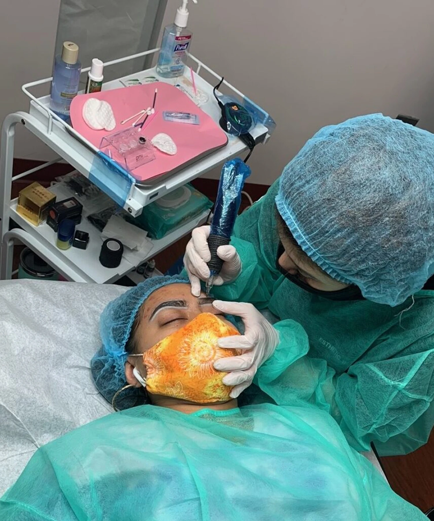 A person lying on a medical bed receives a facial procedure from a professional wearing surgical attire, in a clinical setting with medical equipment nearby.