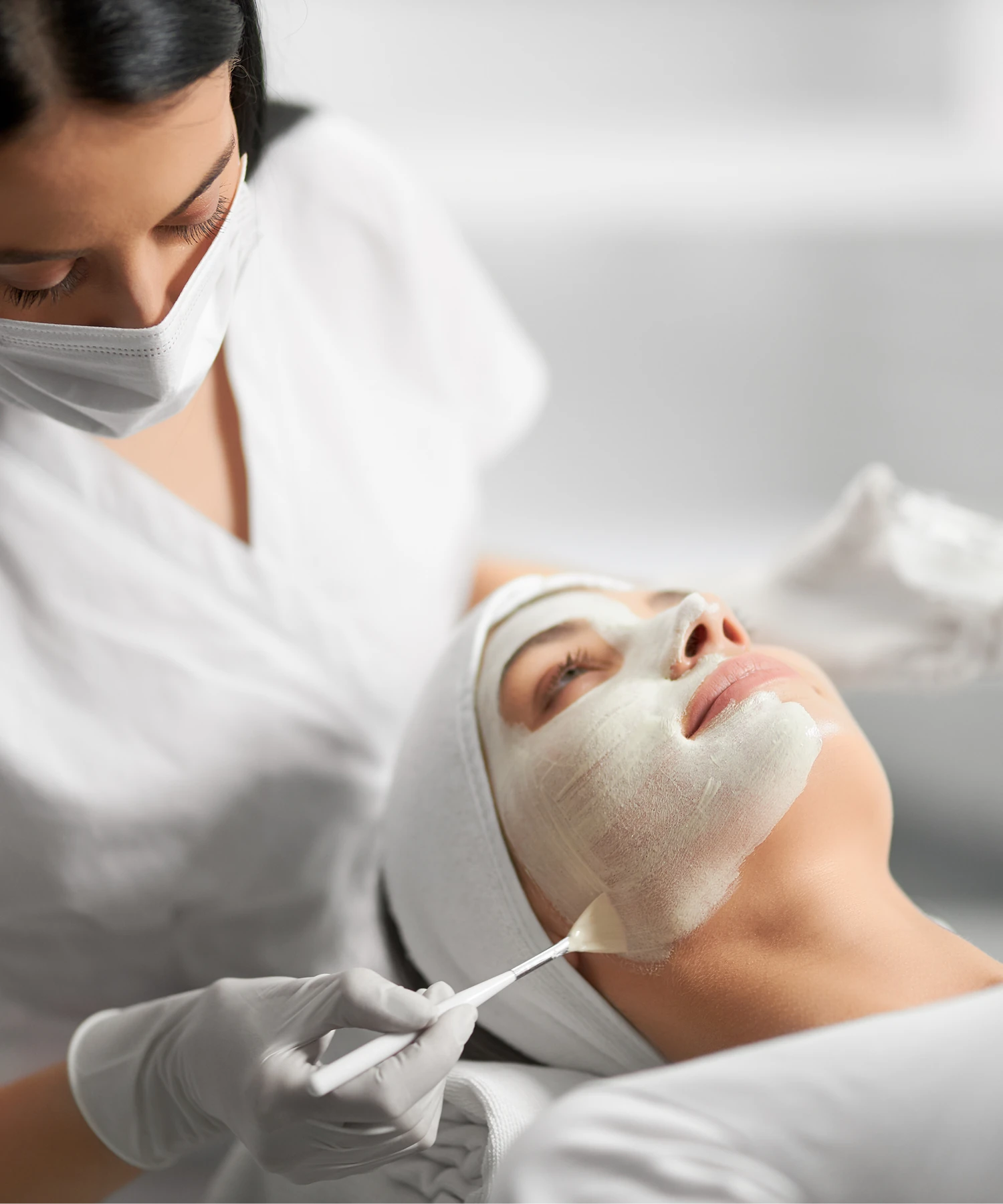 A woman undergoes a facial treatment with a white mask applied, assisted by a professional wearing gloves and a mask in a spa setting.