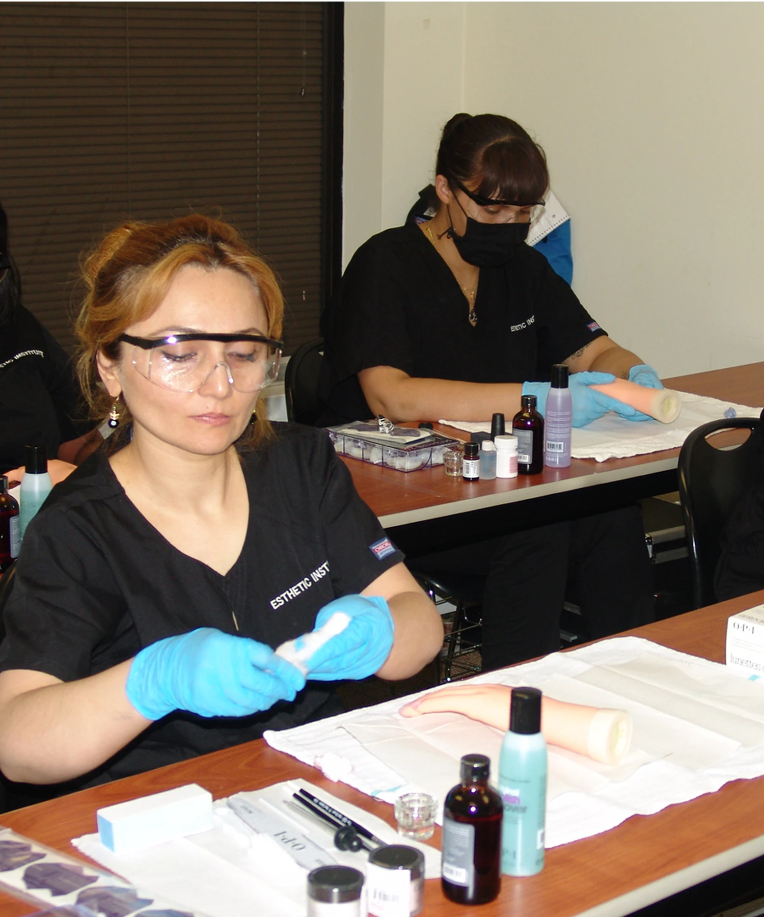 Two technicians in black uniforms and protective eyewear work on beauty applications at a desk, using skincare products and tools, with focus on practical tasks in a classroom setting.