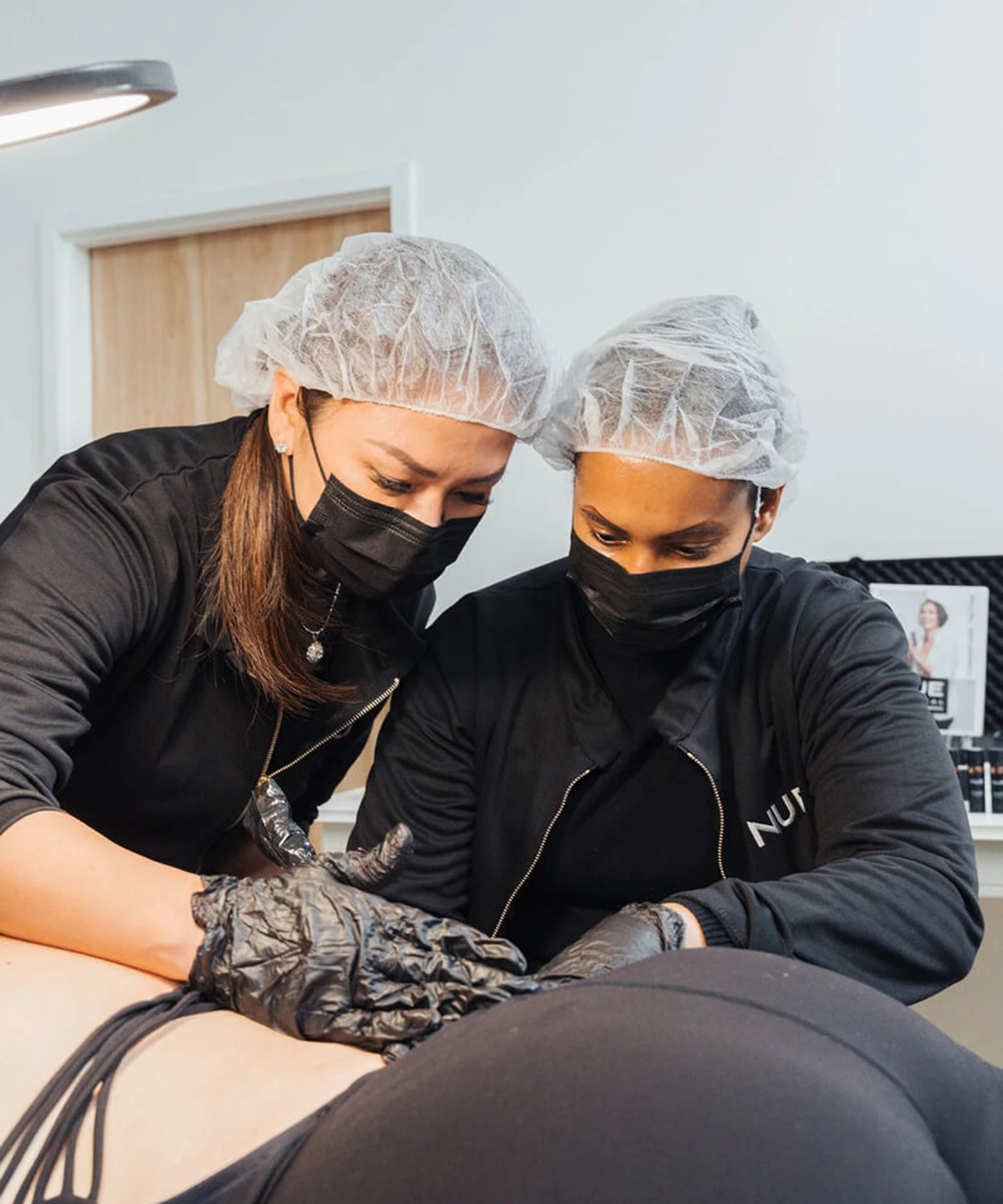 Two professionals wearing masks and hairnets focus on a cosmetic procedure. One holds tools, while the other assists, next to a prone person covered with a cloth.