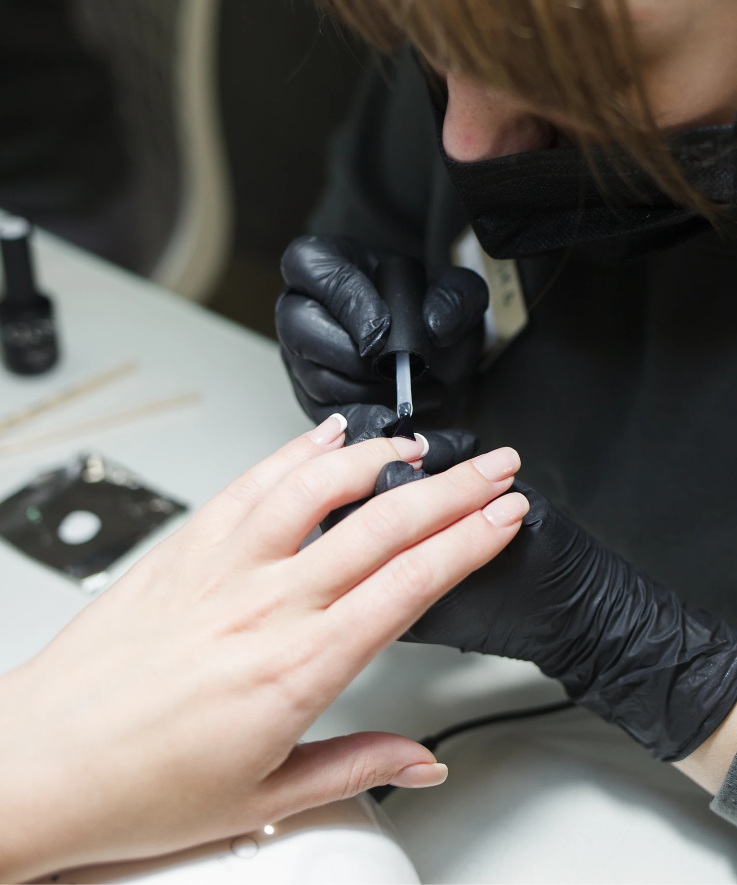 A person applies nail polish to another person's fingernails. Both wear black gloves and the setting appears to be a nail salon.