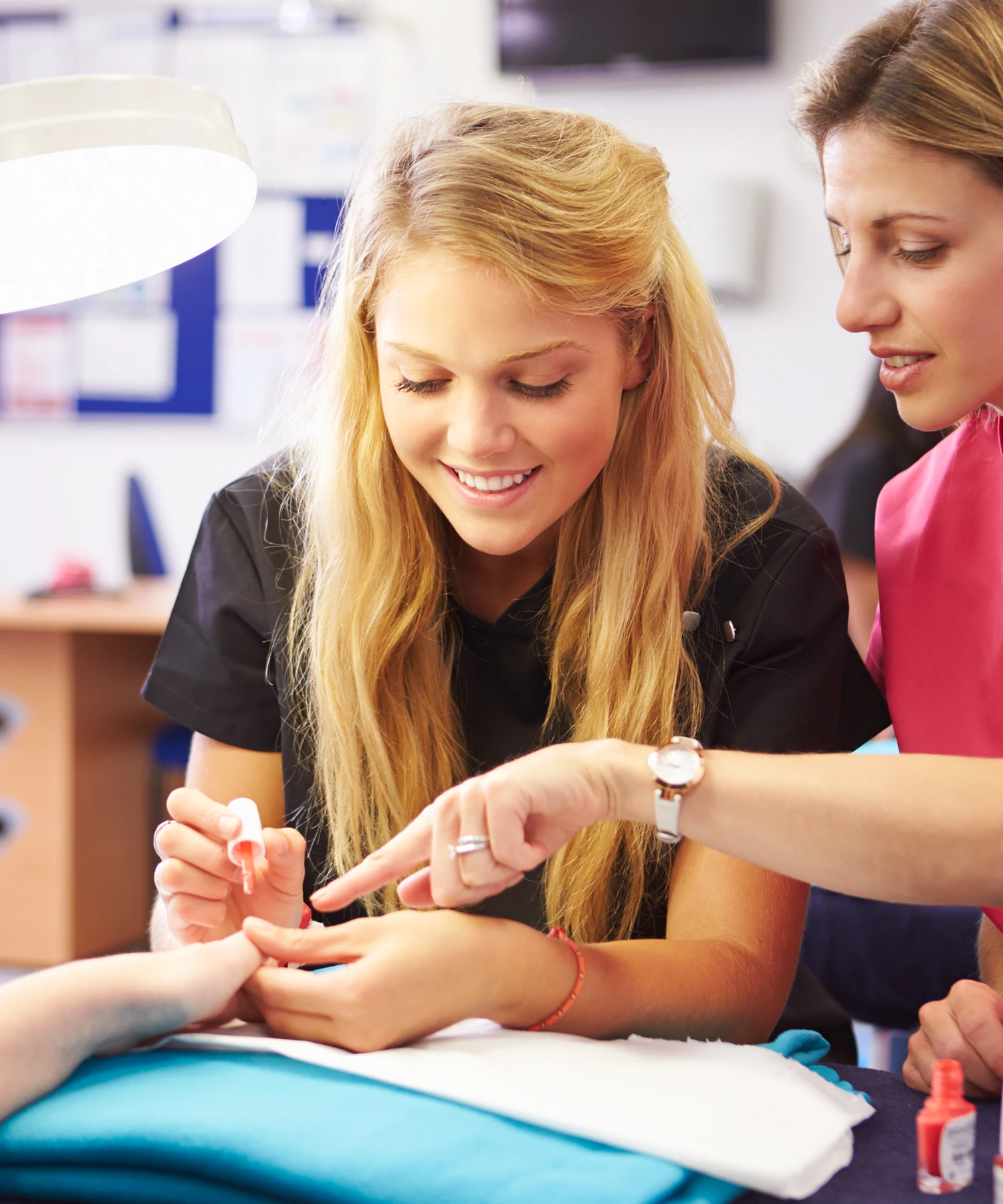 Person learning to apply nail polish while another observes, both focused on a hand resting on a blue towel.