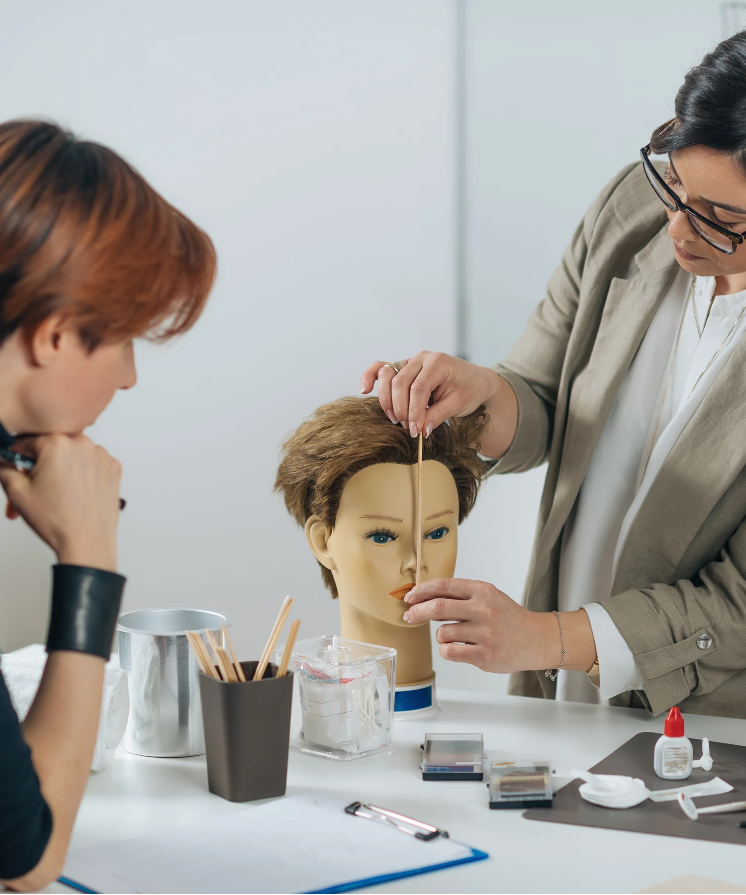 Two people are working on a mannequin head, applying a strip to its face. Various tools are spread on the table.