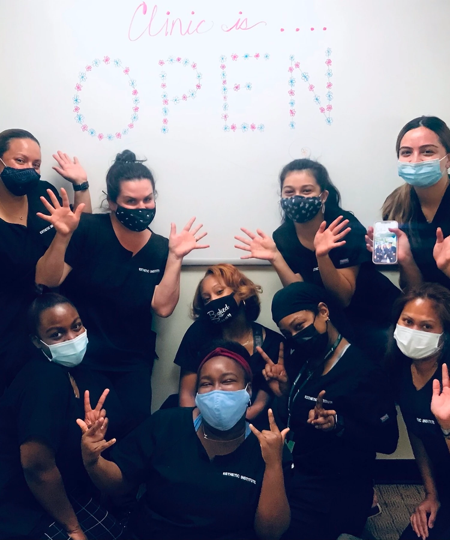 Group of nine people wearing masks and scrubs posing in front of a whiteboard with "Clinic is Open" written on it.
