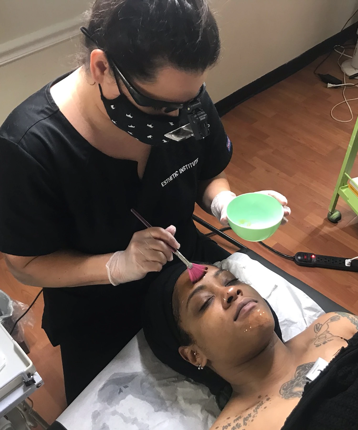A facial treatment is being applied to a person lying down, with a professional wearing gloves and a mask using a brush.