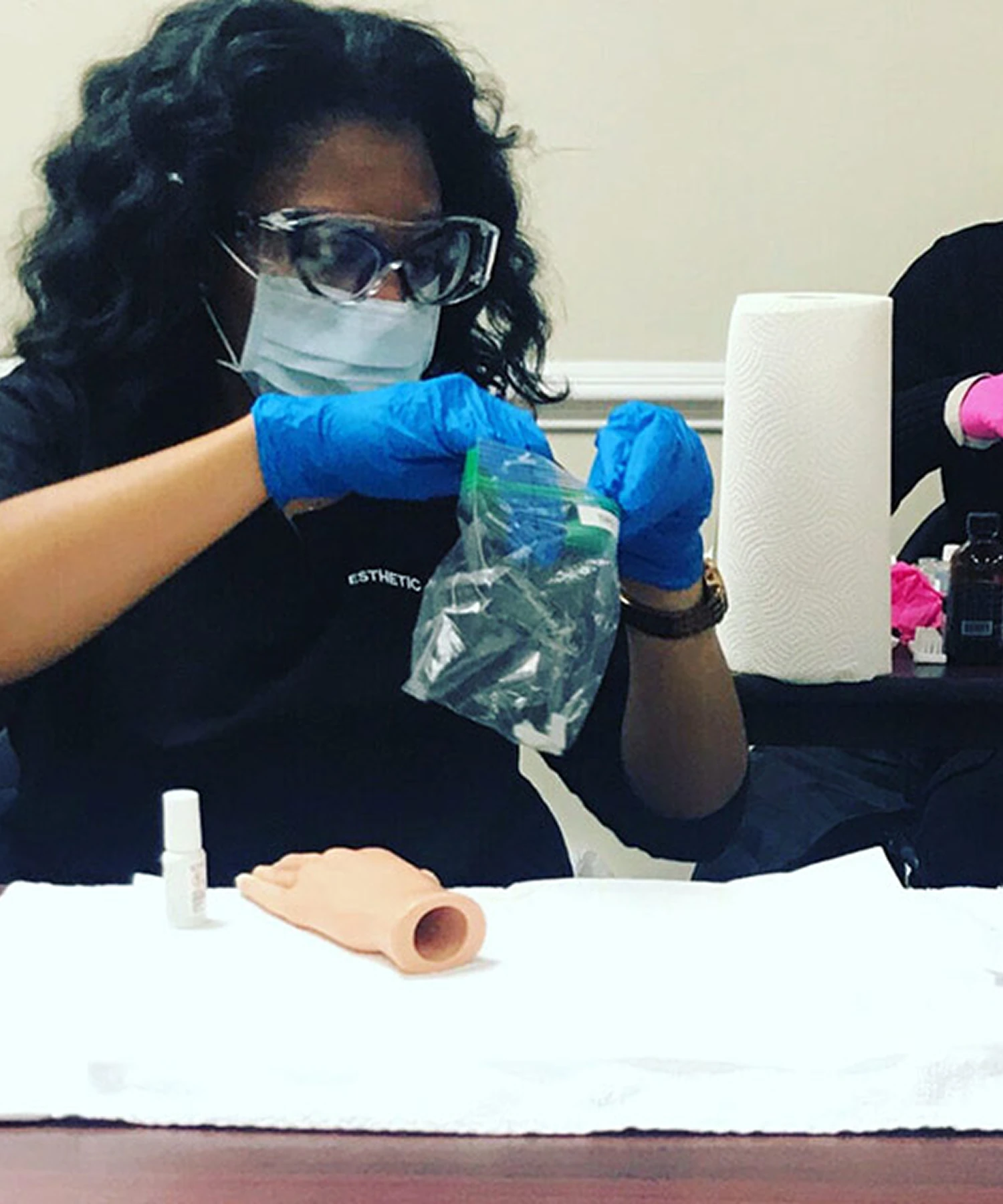 Person wearing goggles, mask, and gloves works with tools from a small bag at a table. A practice hand model, paper towel roll, and small bottle are also visible.