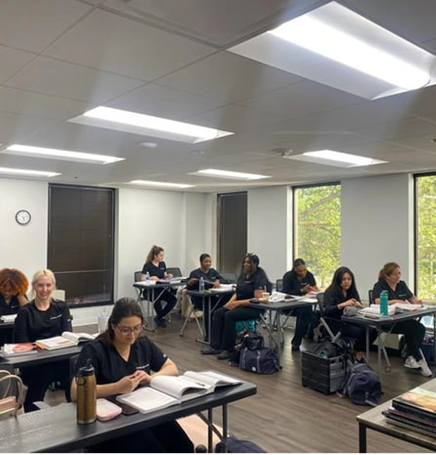Students sitting at desks in a classroom with notebooks and backpacks, some are writing while others are looking ahead. Bright overhead lights illuminate the room.