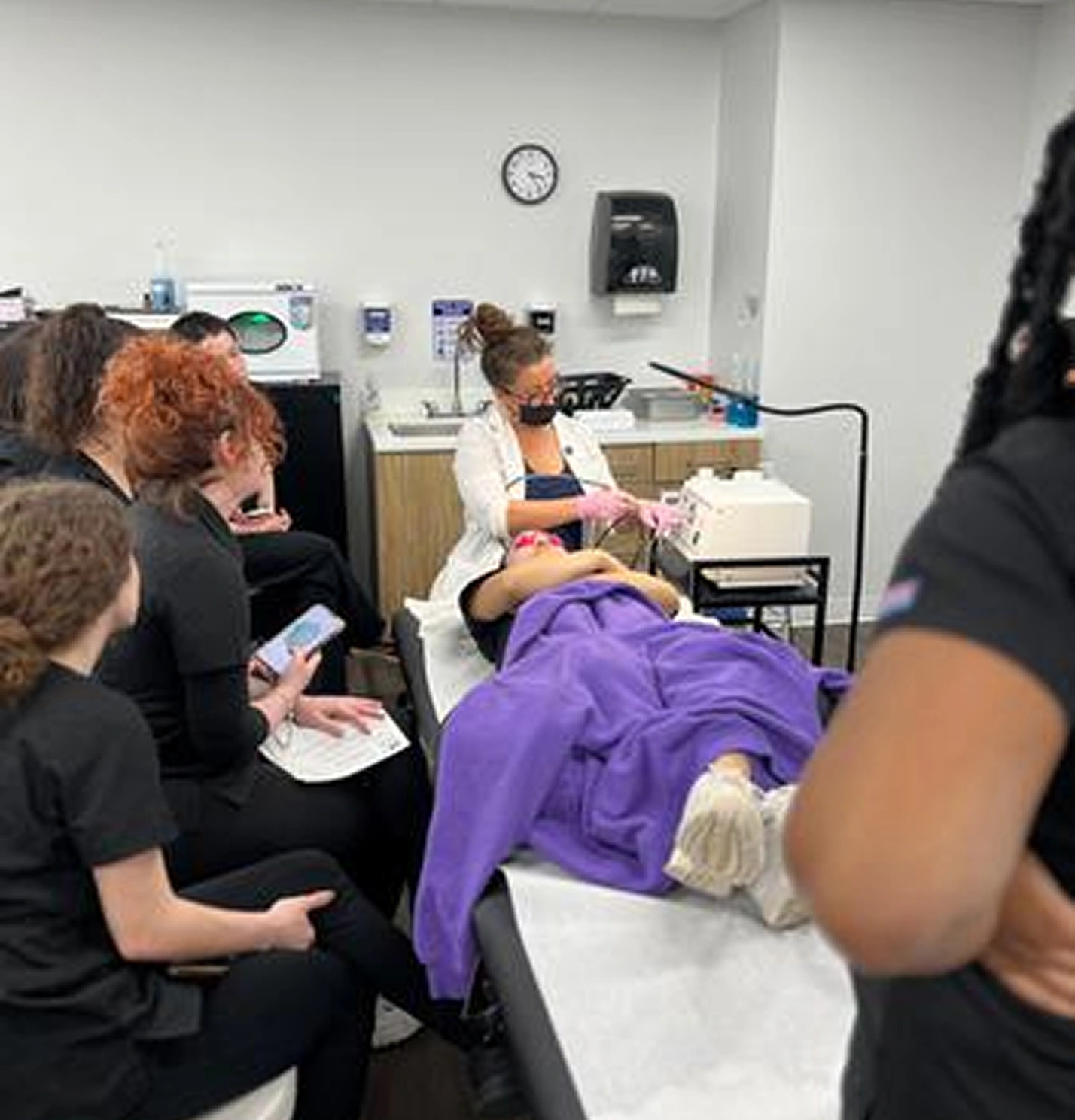 A person in a salon chair receives a skincare treatment under a purple blanket, surrounded by multiple observers in a clinic setting.