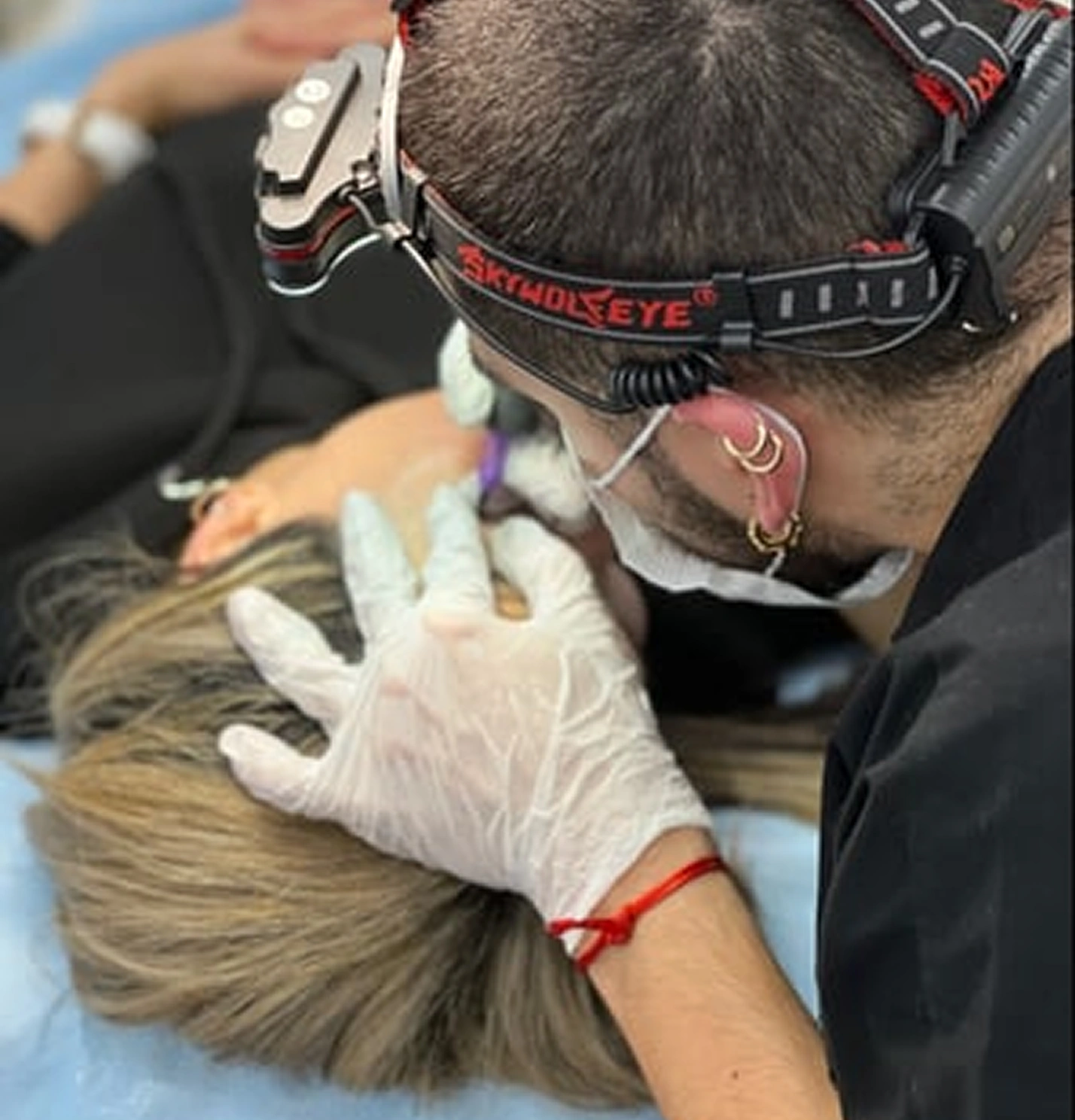 A person wearing magnifying glasses and gloves works on a client's lips, applying a cosmetic or medical procedure. The client is lying down with a cloth over their clothing.
