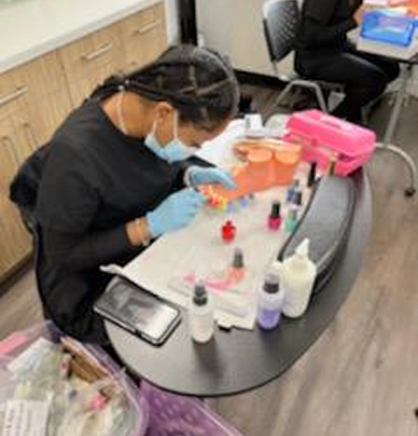 Person with braided hair, wearing a mask and gloves, painting artificial nails at a small table with nail polish and supplies.