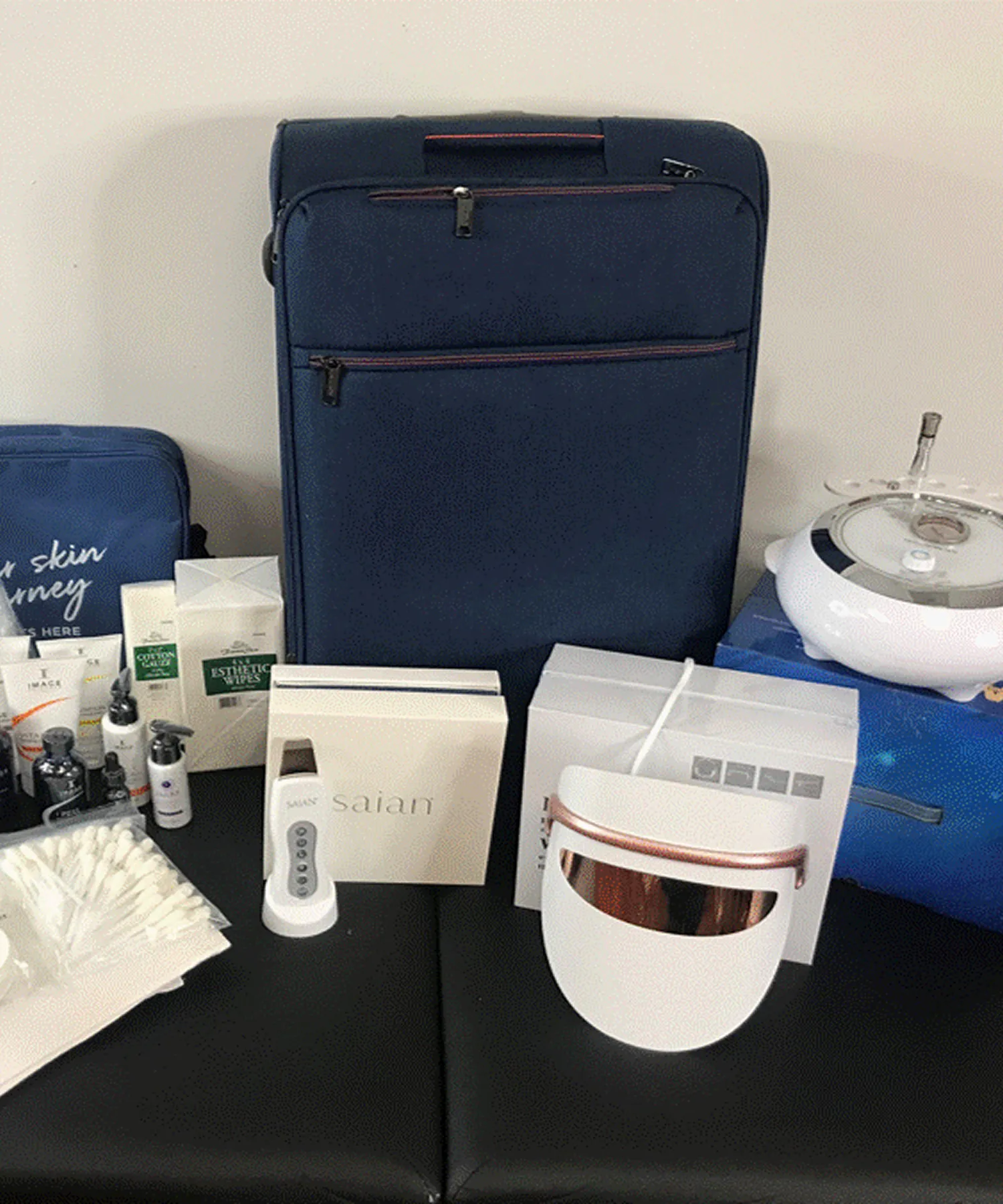 A blue suitcase surrounded by beauty products, including a white LED face mask, creams, and cosmetic tools, displayed on a black surface.