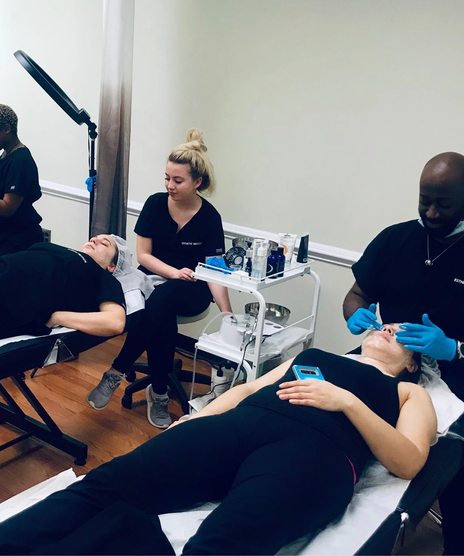 Two people receive facial treatments while lying on beds. Two technicians, one each per client, work on their treatments. The clients are dressed in black, with one holding a blue phone.