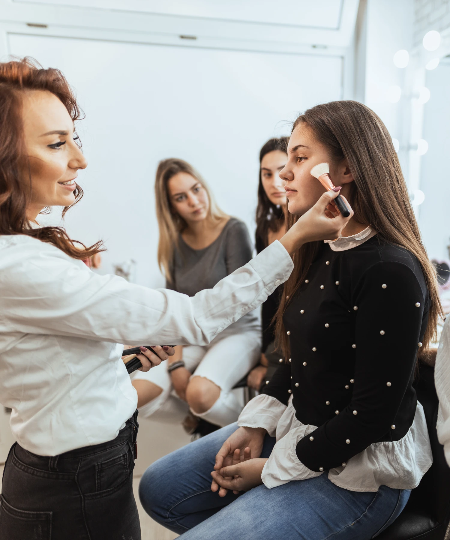 A woman applies makeup with a brush to another woman's face in a studio, while two others watch intently in the background.