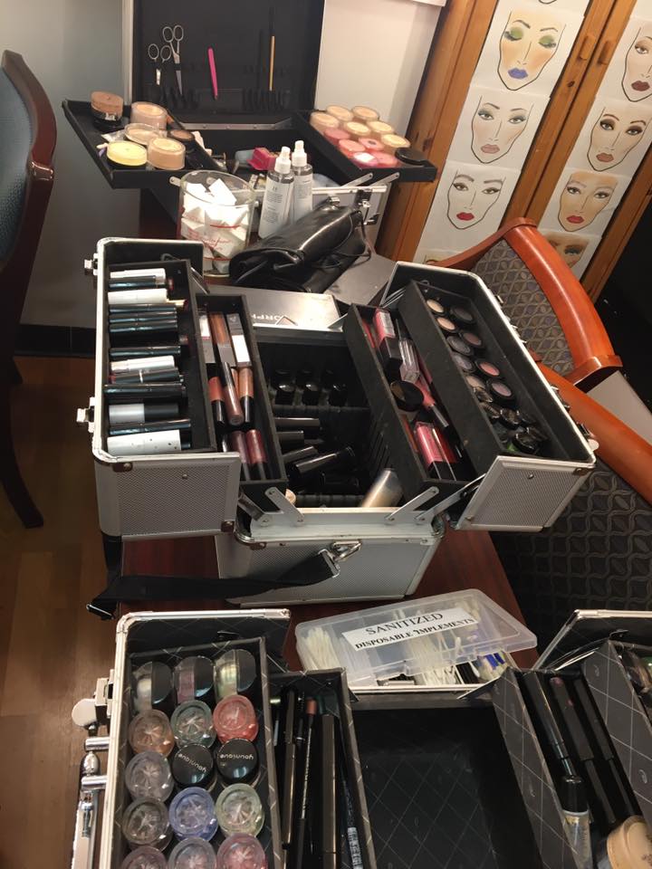 A makeup station with open cases displaying various cosmetics, brushes, powders, and lipsticks, set on a wooden-floored room.